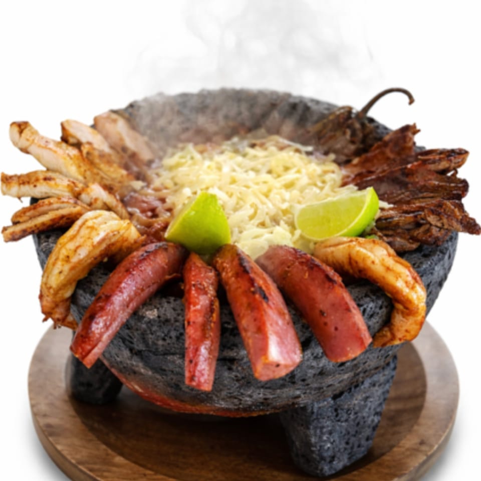 Steaming molcajete filled with grilled chicken, steak, sausage, shrimp, melted cheese, and lime wedges.