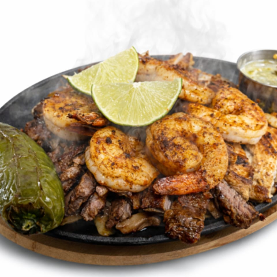 Sizzling fajitas with steak, shrimp, chicken, lime wedges, and a green pepper on a dark platter.