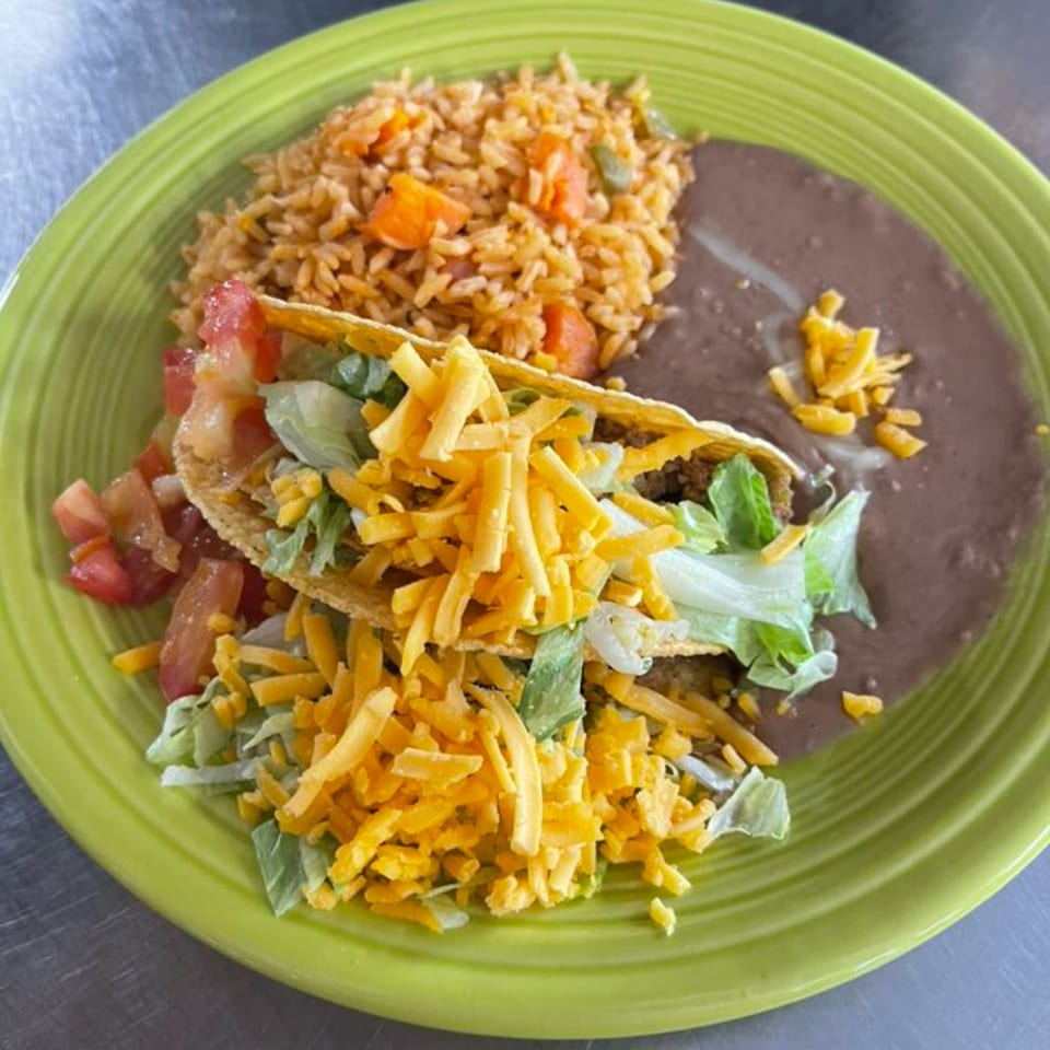 Two crispy tacos with lettuce, cheese, and tomatoes, served with rice and refried beans on a green plate.