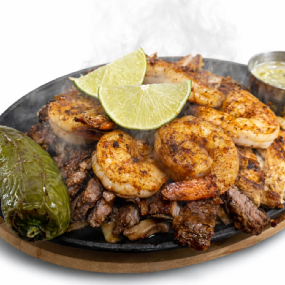 Sizzling fajitas with shrimp, steak, chicken, lime wedges, and a green pepper on a hot plate.