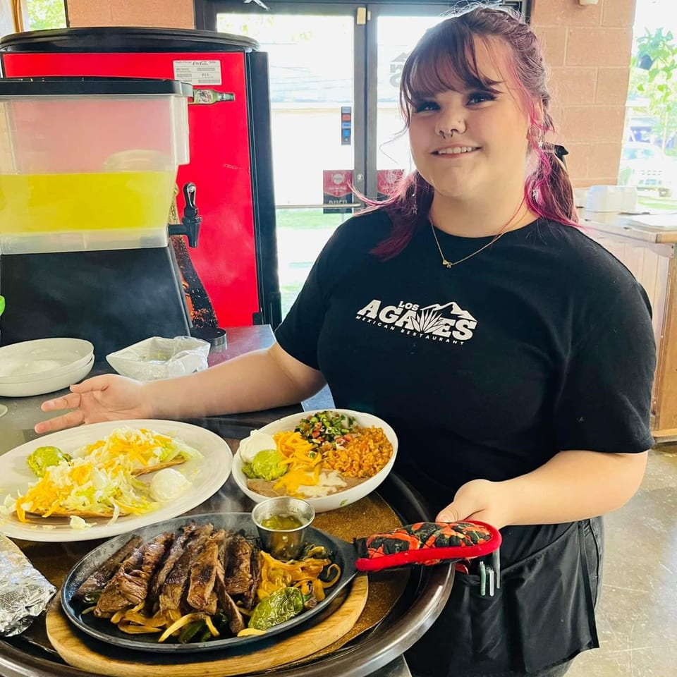 A smiling Los Agaves server holds a tray with sizzling fajitas, tacos, and a rice and bean bowl.
